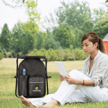 Cooler backpack chair sitting in the grass next to a woman reading. 