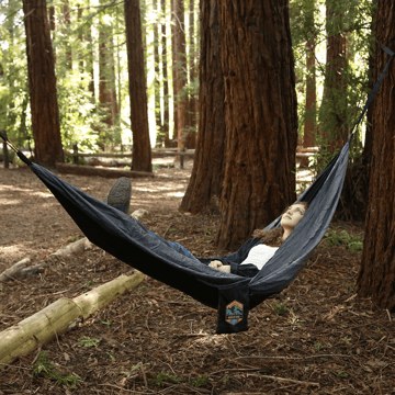 Woman laying in a hammock strung between 2 trees in a wooded area. 